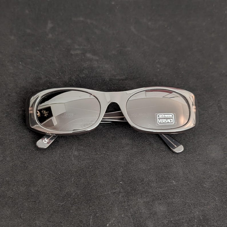 Puede incluir: Gafas de sol Versace grises oscuras con lentes ovaladas. Las gafas tienen una montura gruesa y lentes oscuras. El logotipo de Versace es visible en las lentes y las patillas. Las gafas est&aacute;n sobre una superficie negra.