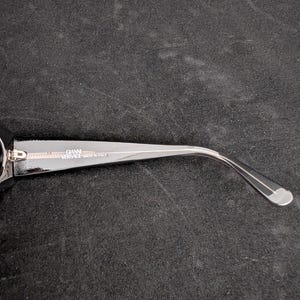 Puede incluir: Primer plano de una patilla de gafas negra con el texto "GIANNI VERSACE MADE IN ITALY" impreso en blanco. La patilla es curva y tiene una punta plateada. El fondo es una superficie oscura y texturizada.