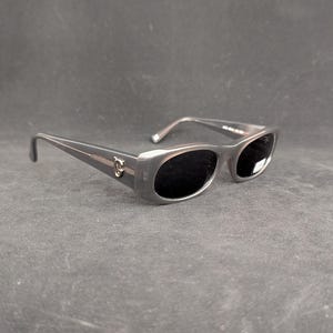 Puede incluir: Gafas de sol grises con lentes oscuras. Las gafas tienen una montura rectangular con un peque&ntilde;o emblema dorado en el lateral. Las patillas son finas y se curvan hacia la oreja.