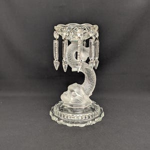 May include: A clear glass candle holder featuring a dolphin design. The base is round with a decorative edge, and the dolphin's tail supports a bowl with hanging crystal prisms. The dolphin's head is at the base of the holder.