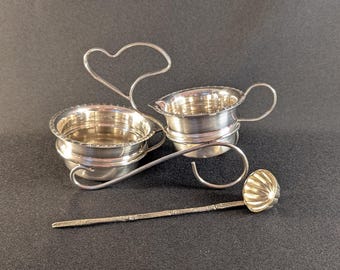 May include: A silver creamer and sugar bowl set with a heart-shaped handle. The set includes a small creamer with a handle, a sugar bowl, and a small spoon. The pieces are made of shiny silver metal and sit on a dark surface.