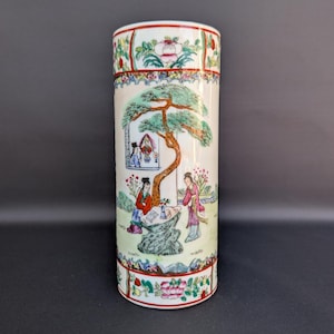 Può includere: Un vaso cilindrico in porcellana con una scena dipinta a mano di figure in un giardino con un grande pino. Il design include bordi floreali decorativi in rosso, verde e rosa sui bordi superiore e inferiore del contenitore di colore bianco.