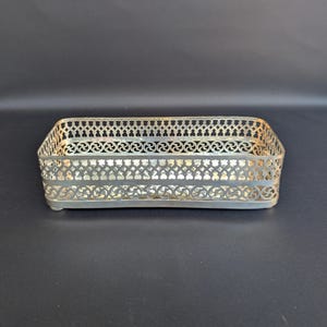 Könnte beinhalten: Ein rechteckiges Tablett aus silberfarbenem Metall mit einem komplizierten, durchbrochenen Gitterdesign an den Seiten. Die Struktur weist ein sich wiederholendes geometrisches Muster aus kleinen Ausschnitten und Kreisen auf.