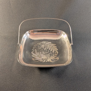 Peut inclure: Un plat de service rectangulaire argenté avec une poignée. Le plat présente un motif floral en relief au centre. La poignée est arquée et fixée sur les côtés du plat. Le plat est sur une surface sombre.