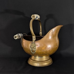 May include: An antique copper coal scuttle with a brass base and handle. The handle features white and blue ceramic detailing. A brass lion's head decorates the front. The scuttle has a spout and a dark interior. A vintage decorative item.
