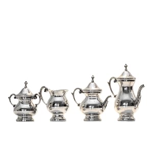 Könnte beinhalten: Ein silbernes Teeservice mit Teekanne, Sahnekännchen, Zuckerdose und einer kleinen Schale mit Deckel. Das Set hat verzierte Griffe und Deckel, die das Licht reflektieren. Die Teile sind vor weißem Hintergrund angeordnet.