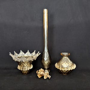 Peut inclure: Ensemble de vases et de bols décoratifs en verre dans des tons or et marron. La collection comprend un vase long et élancé, un bol avec un bord en forme de couronne et un petit vase rond avec une base décorative. Un élément floral doré est également présent.
