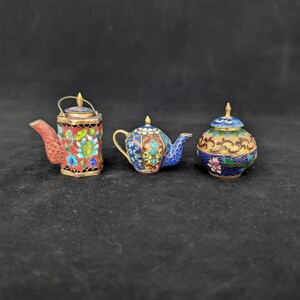 Pode incluir: Três recipientes decorativos em miniatura com um intrincado trabalho de esmalte cloisonné em tons vibrantes de vermelho, azul, verde e dourado. O conjunto inclui um bule hexagonal com pega, um pequeno bule redondo e um frasco com tampa e motivos florais.