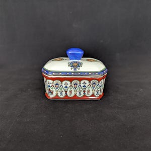 May include: A rectangular ceramic trinket box with a lid. The box has a white top with floral and blue designs, a red base with a repeating pattern, and a blue knob on the lid. The box is on a dark background.
