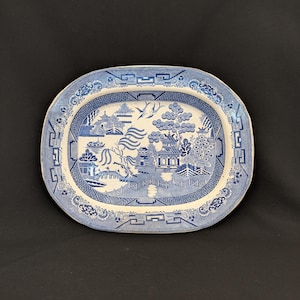 Op de afbeelding: Ovale blauwe en witte keramische serveerschaal met een gedetailleerd Willow-patroon. De schaal heeft een centrale landschapsweergave met pagodes, bruggen en bomen, omgeven door een decoratieve rand. Een klassiek stuk om eten te serveren.