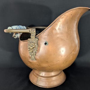 Könnte beinhalten: Ein kupferner Kohleeimer mit einem dekorativen Löwenkopf aus Messing und einem Griff mit blau-weißem Porzellangriff. Der Eimer hat einen abgerundeten Körper und eine schnabelartige Öffnung und steht auf einem Sockel.