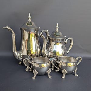 Puede incluir: Un juego de té de cuatro piezas de metal plateado que incluye una cafetera alta, una tetera más pequeña, una lechera y un azucarero. Cada pieza tiene una superficie pulida y reflectante, asas curvas ornamentadas y pies decorativos.