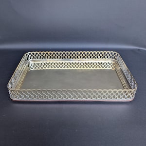 Könnte beinhalten: Ein rechteckiges Tablett aus silberfarbenem Metall mit einer polierten, reflektierenden Basis und einem erhöhten, filigranen Gitterrand mit gewelltem Abschluss. Das Tablett hat eine metallische Oberfläche und ein strukturiertes Design.
