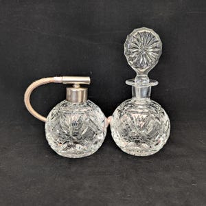 Peut inclure: Deux flacons de parfum en verre taillé transparent. L'un a une forme bulbeuse avec un dessus en métal argenté et un atomiseur rose. L'autre flacon a un bouchon en verre taillé assorti. Les deux flacons ont un motif détaillé en forme d'éclat d'étoile.