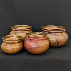 May include: Four decorative, round pots with a metallic bronze finish and gold rims. The pots vary in size, with a reddish-brown hue on the smallest pot. The background is a solid black.