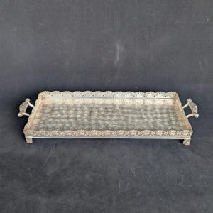 Può includere: Un vassoio da portata rettangolare in metallo color argento caratterizzato da un bordo smerlato decorativo con un motivo ripetitivo. Il vassoio presenta due manici ornati sui lati e poggia su quattro piedini, con una superficie testurizzata.