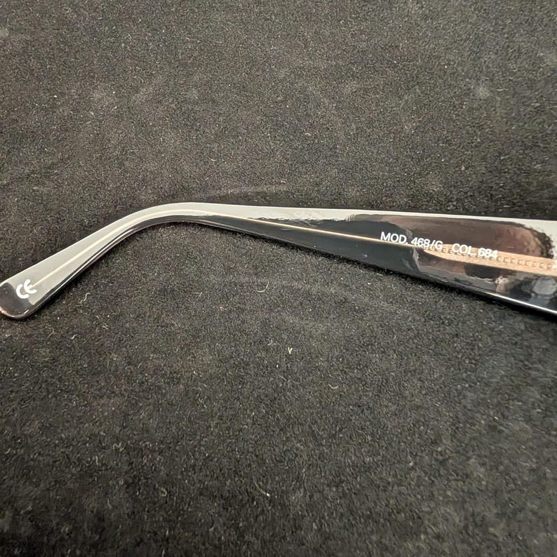 Puede incluir: Primer plano de una patilla de gafas plateada y negra. La patilla tiene el texto "MOD 468/G COL 684" impreso. La patilla es curva y tiene una marca CE.