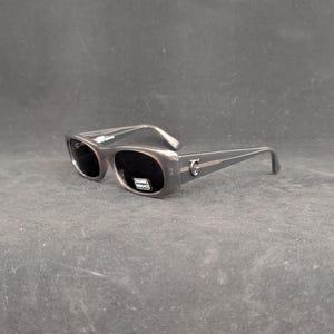 Puede incluir: Gafas de sol grises con lentes oscuras. Las gafas tienen una montura rectangular y un acento plateado en las patillas. El nombre de la marca es visible en la lente.