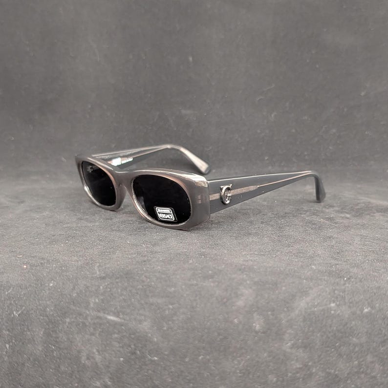 Puede incluir: Gafas de sol grises con lentes oscuras. Las gafas tienen una montura rectangular y un acento plateado en las patillas. El nombre de la marca es visible en la lente.