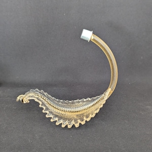 Può includere: Un oggetto decorativo e curvo con un design strutturato a forma di foglia. L'oggetto è principalmente dorato con un tappo in metallo argentato nella parte superiore. La base dell'oggetto ha un bordo seghettato dettagliato.