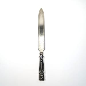 May include: A silver letter opener with a pointed blade and ornate handle. The blade has etched detailing near the tip. The handle features a decorative design with a heart-shaped element and a detailed base.