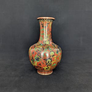 Op de afbeelding: Een bolvormige vaas met een smalle hals en uitlopende rand, voorzien van een dicht, ingewikkeld patroon van gestileerde bloemen en bladeren in levendige tinten rood, groen, zwart en goud, afgewerkt met een koperkleurige rand en voet.