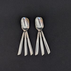 Può includere: Due set di cucchiai d'argento su sfondo nero. Ogni set ha tre cucchiai con manici decorati. I cucchiai hanno una superficie lucida e i manici presentano un elaborato motivo floreale.