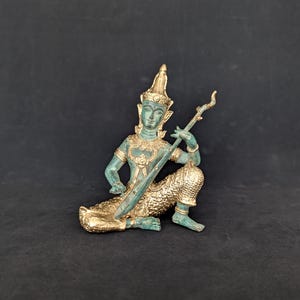Op de afbeelding: Een metalen beeldje van een zittende godheid die een snaarinstrument bespeelt. Het figuur heeft een verdigris groene patina op de romp en het gezicht, gecontrasteerd met gepolijste goudkleurige accenten op de kroon, sieraden en onderkleding.