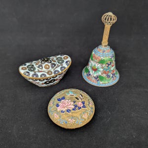 Pode incluir: Uma coleção de três objetos decorativos: uma caixa cloisonné em forma de barco com padrão de moedas, um sino com design floral em esmalte e cabo dourado, e um estojo redondo de latão com um motivo de rosa rosa e borboletas em esmalte.