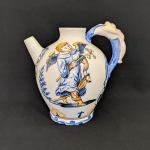 Può includere: Brocca in ceramica bianca con beccuccio e manico, con un disegno dipinto a mano di un angelo in abito blu che porta una croce e un cavallo. La brocca ha dettagli blu e gialli.