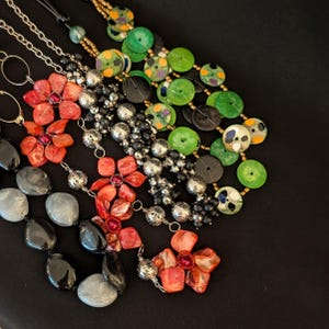 May include: A selection of handmade necklaces in diverse styles and colours. The necklaces feature flower-shaped pendants in red and orange, and beaded strands in black, silver, and green. The necklaces are presented on a black surface.