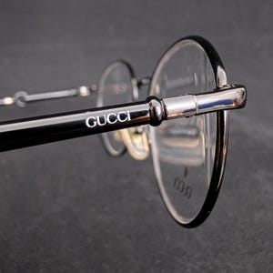 Op de afbeelding: Close-up van een Gucci-bril. De bril heeft een zwart montuur met het Gucci-logo in wit op de poot. De glazen zijn helder en rond, met een zilveren metalen brug.