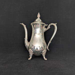 Puede incluir: Una cafetera de metal plateado pulido con un cuerpo alto y esbelto, un pico curvo y un asa ornamentada. La cafetera se apoya sobre tres patas decorativas e incluye una tapa a juego con un remate puntiagudo en la parte superior del recipiente.