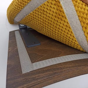 May include: A dark brown wooden shelf with a black metal L-bracket attached to the wall. A textured mustard yellow fabric pad with grey hook-and-loop fastener strips is being positioned onto the matching grey strips on the wooden shelf surface.