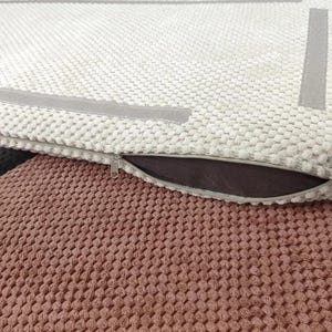 May include: A rectangular cushion cover featuring a cream-colored textured popcorn knit fabric with smooth grey border strips. The cover includes a metal zipper closure along the side, revealing a dark brown interior lining against a dusty rose base.