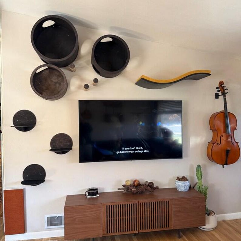 May include: A wall-mounted cat furniture set featuring two large dark circular pods, three small circular shelves, and one long curved yellow and dark shelf. A flat-screen television is mounted centrally below the pods, above a wooden media console.