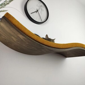 May include: A curved, wall-mounted wooden cat shelf featuring a dark brown wood grain finish and a textured, mustard yellow fabric cushion. The shelf has a wave-like design and is mounted horizontally on a white wall.