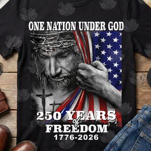 May include: Black t-shirt featuring a graphic of Jesus holding an American flag. The text reads "ONE NATION UNDER GOD" above the image and "250 YEARS of FREEDOM 1776-2026" below. The design is patriotic and religious.
