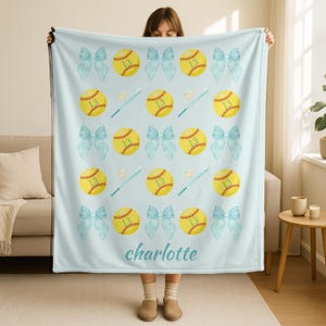 May include: A pale blue blanket with a repeating pattern of yellow softballs with the number 13, light blue bows, and baseball bats. The name "charlotte" is printed in a script font at the bottom.
