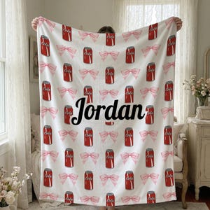 May include: White blanket featuring a pattern of red cola cans and pink bows. The name "Jordan" is printed in black script across the centre. A decorative blanket for a bedroom or living space.