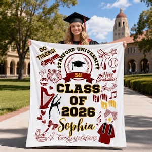 Könnte beinhalten: Eine weiße Abschlussdecke mit weinroten und goldenen Grafiken. Die Decke enthält den Text "Stanford University", "Class of 2026" und "Sophia Congratulations". Dekorative Elemente sind ein Tiger, Baseball, Noten und eine Abschlusskappe.