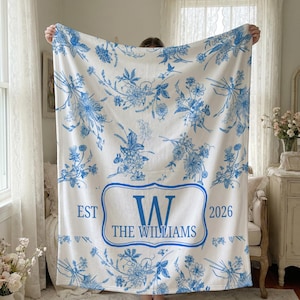 May include: White blanket with a blue floral design. The blanket features a central emblem with the letter "W" and the words "THE WILLIAMS" below it. The blanket also has the text "EST 2026".