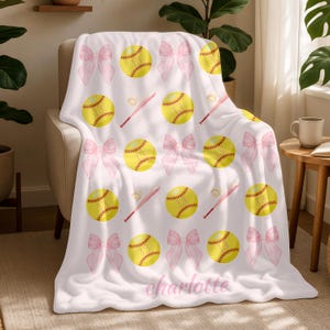 May include: A white blanket with a softball design. It features yellow softballs, pink bows, and pink baseball bats. The name "charlotte" is printed on the blanket in a cursive font.