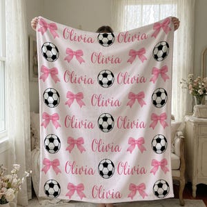 May include: A light pink blanket featuring a repeating pattern of soccer balls, pink bows, and the name "Olivia" in a script font. The soccer balls are black and white.