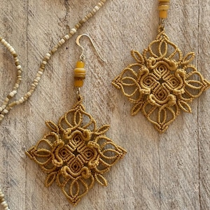 May include: A pair of handcrafted macrame earrings in a geometric design. The earrings are a warm, golden-brown colour and feature a square shape with intricate knotting. A beaded necklace in neutral tones is also visible.