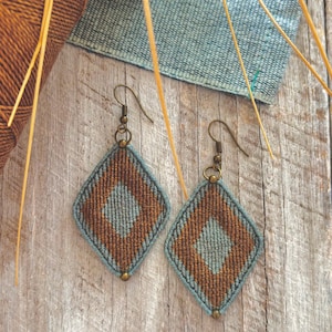 May include: A pair of diamond-shaped macrame earrings in brown and blue, with bronze-coloured ear wires. The earrings are displayed on a wooden surface, alongside spools of thread. The earrings are a handcrafted item.