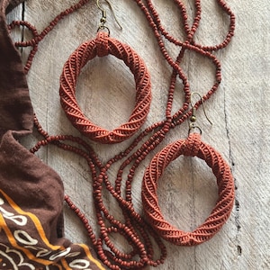 May include: A set of two circular macrame earrings in a terracotta color with bronze-toned metal hooks, accompanied by a long necklace made of small terracotta-colored beads and a brown fabric cloth featuring a yellow and white floral pattern.
