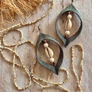 May include: A pair of olive green fabric earrings featuring a central cowrie shell, small white porous beads, and brown wooden spacers, accompanied by a long necklace made of small, light-colored seed beads with a metallic hook closure.