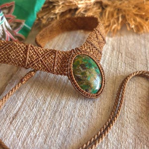 May include: A brown macrame necklace featuring an oval, green and brown stone pendant. The necklace has a woven design and a long, braided cord. The background includes a green and red patterned fabric and a wooden surface.