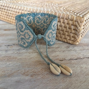 May include: A wide woven bracelet featuring a teal and beige geometric pattern. The bracelet has a drawstring closure with two thin cords, each ending in a small, smooth, cream-colored cowrie shell. The design is symmetrical and flat.